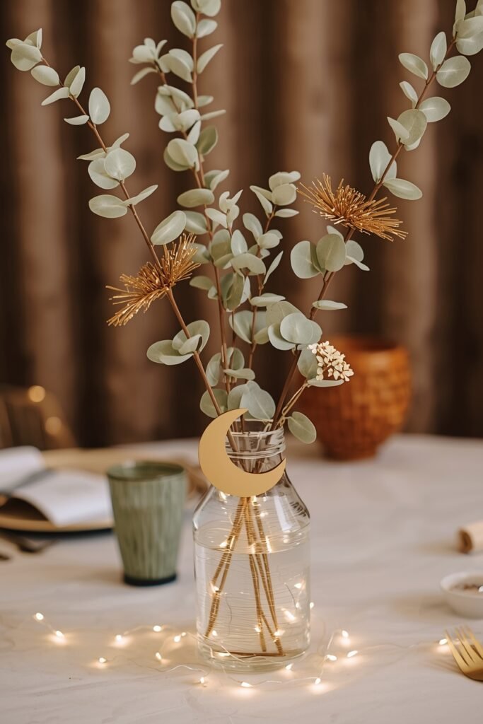 A simple DIY Eid table centrepiece — a glass vase with faux eucalyptus stems, gold faux flowers and a crescent moon decoration, battery fairy lights coiled at the base, warm golden tones, elegant and affordable
